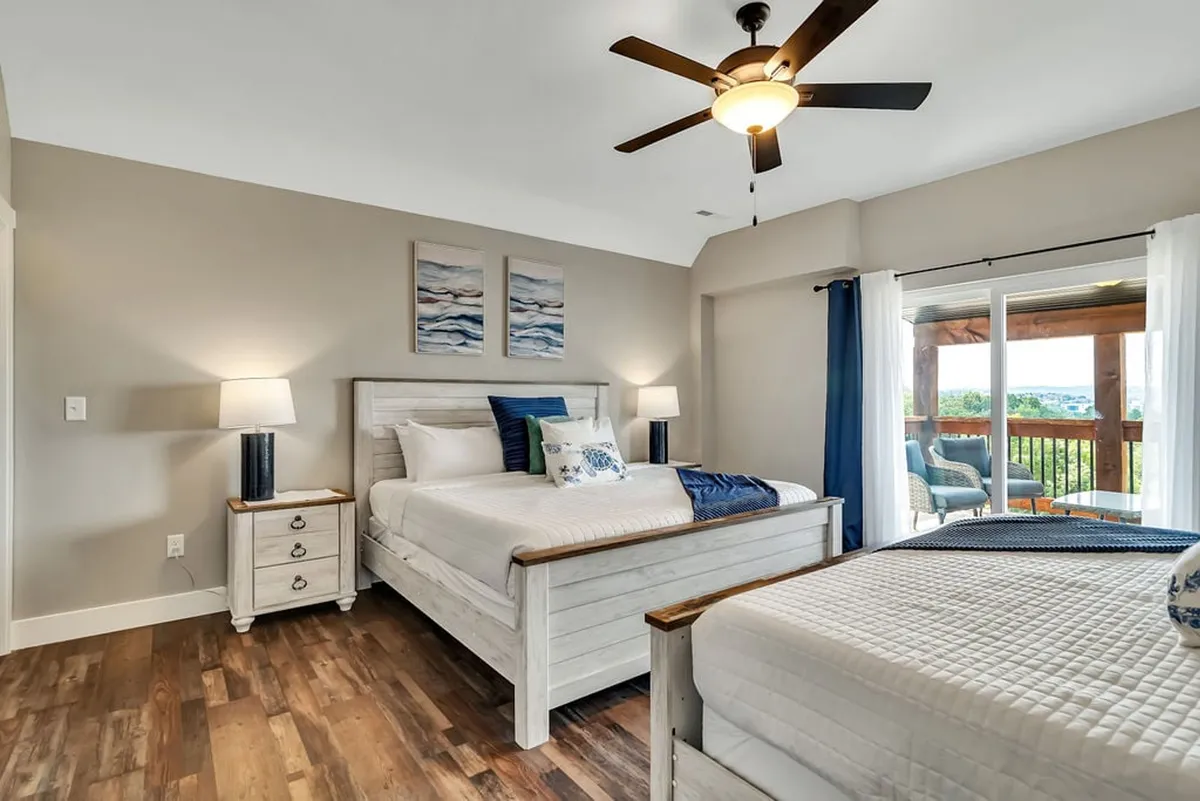 A serene bedroom with two beds, a nightstand with lamps, and tasteful wall art. Large windows allow for natural light, and a balcony is visible, suggesting private outdoor access and a possible lake view.
