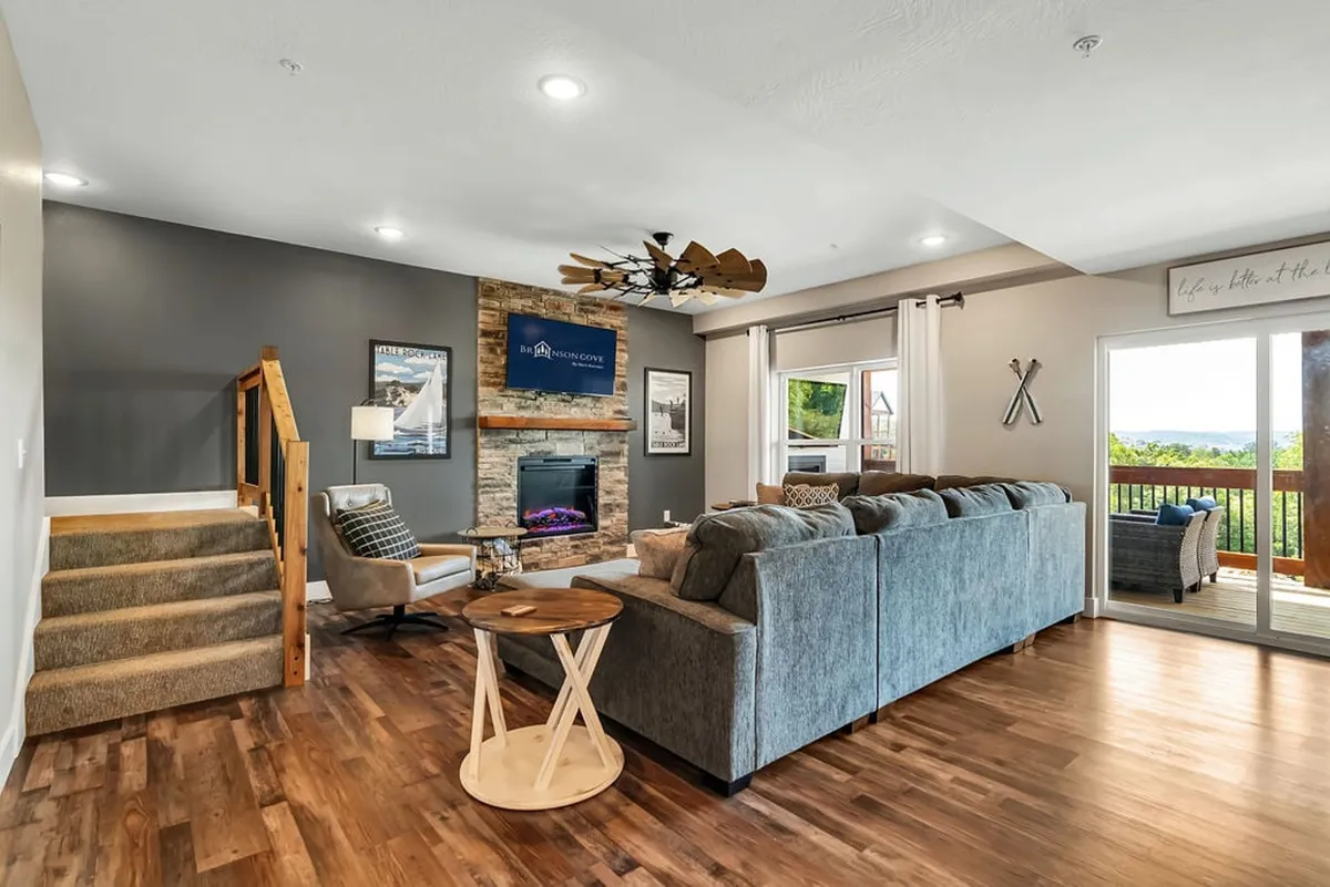 A luminous living area with a large sectional sofa, electric fireplace, wall-mounted television, and expansive windows providing scenic views. The fireplace adds a cozy, intimate feel, while the arrangement encourages conversation and relaxation.