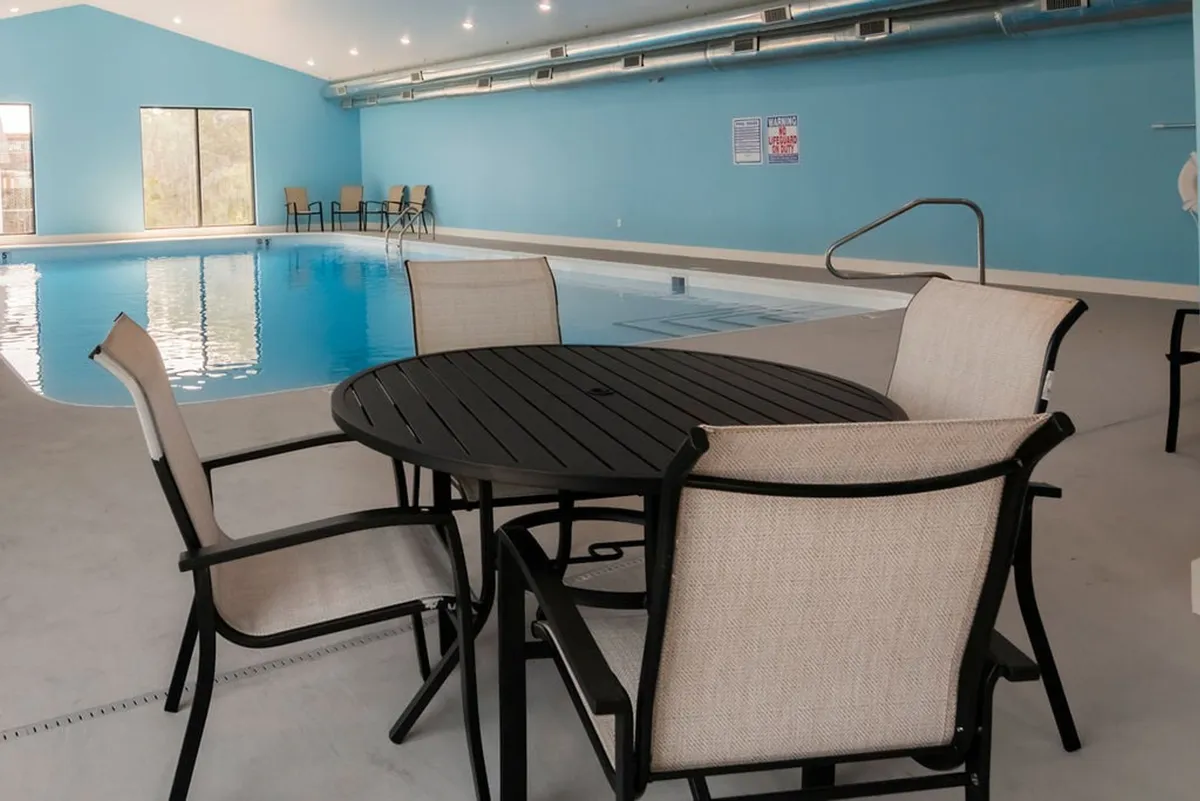 An inviting indoor swimming pool area featuring a tranquil pool, a round table with chairs for relaxation or socializing, and ample lighting that enhances the serene and accessible ambiance. The pool is level with the surrounding space, suggesting ease of entry for guests with mobility considerations.