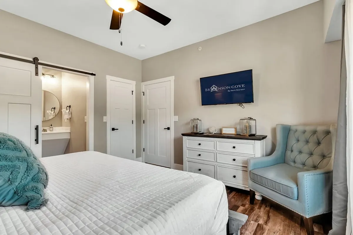 A cozy bedroom with a bed dressed in luxurious white linens and a turquoise throw, a blue armchair, a wall-mounted TV, and large windows that let in the morning sun. The room balances comfort and practicality, suitable for any generation.