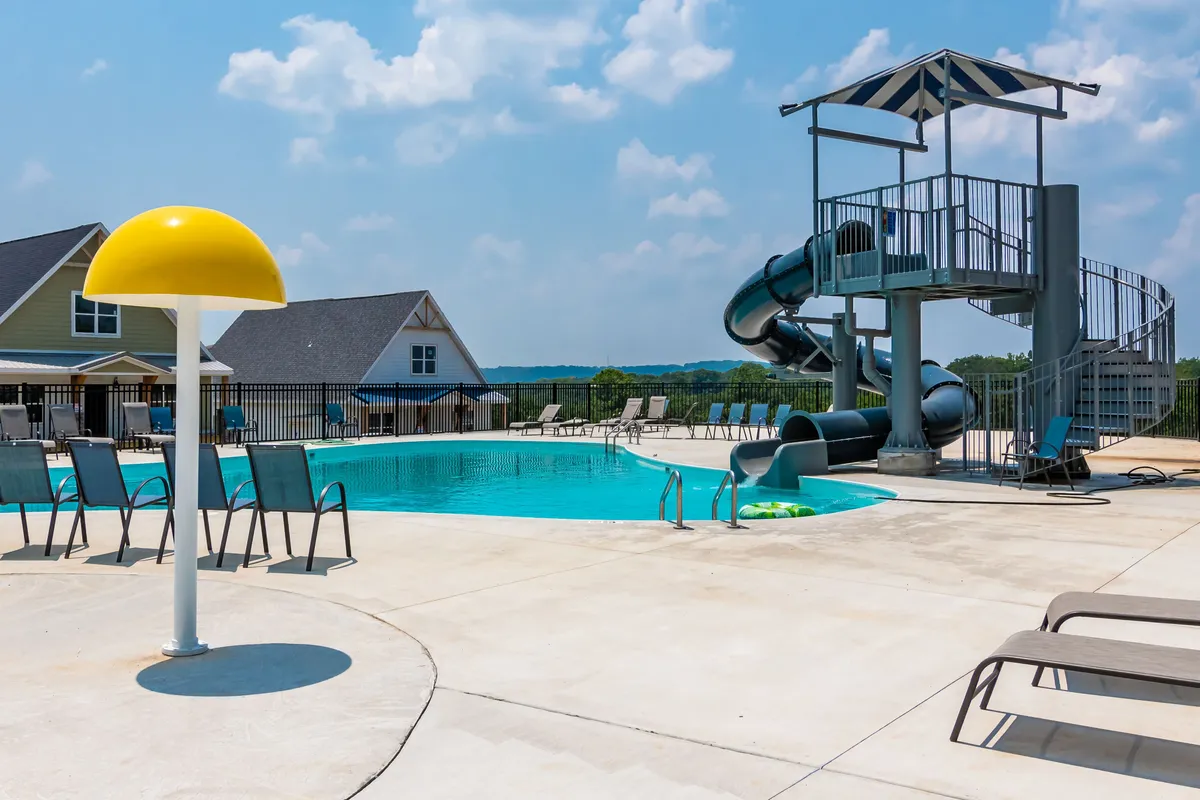 This vibrant exterior image captures the lively outdoor resort-style pool at Branson Cove, complete with a water slide, yellow umbrella, lounge chairs, and surrounding houses. Families and children are seen enjoying the summer sunshine, swimming, and playing in the pool.