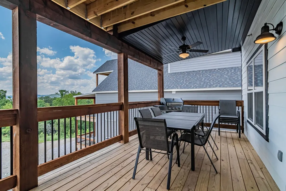 A spacious outdoor dining area with a wooden table set for a group, grill nearby, ceiling fan, and scenic views beyond the railing. The setup invites large groups to dine al fresco and enjoy time together in a casual, relaxed setting.