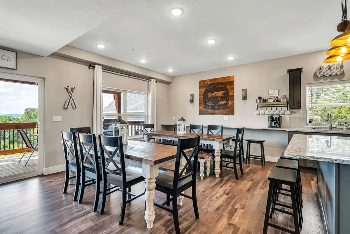 A spacious dining area featuring a large table for group meals, a kitchen bar area with seating, decorative wall art, and a sliding glass door that leads out to a balcony. The setup is ideal for holiday feasts, casual brunches, or sunset dinners with a scenic view.